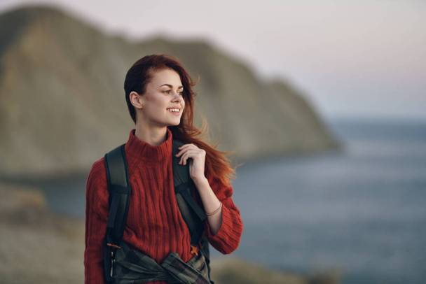 mooie vrouw in een trui houdt een rugzak op haar rug en spreekt in de achtergrond oceaan frisse lucht - Foto, afbeelding