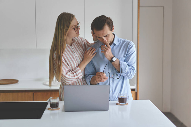 Front view of young couple standing on the kitchen with coffee and laptop - Фото, изображение