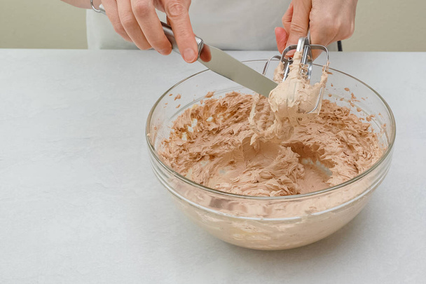 Το γλάσο κέικ σοκολάτας για κέικ σοκολάτας κλείνει σε ένα μπολ. Κρέμα ξυλοδαρμού με ηλεκτρικό μίξερ χειρός, κοντά στο τραπέζι της κουζίνας, γυναικεία χέρια - Φωτογραφία, εικόνα