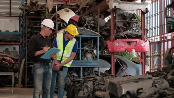 Proprietario caucasico e lavoratore asiatico: Due uomini controllano le parti del motore in un magazzino. Con entrambi che indossano caschi durante l'orario di lavoro, la maggior parte dei prodotti sono macchine nell'industria automobilistica. - Filmati, video