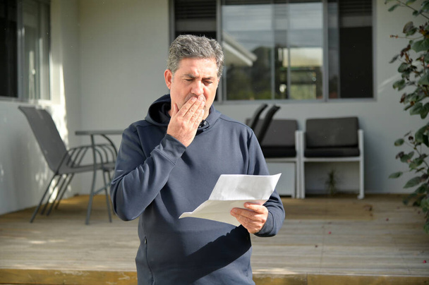 Homme adulte mature choqué (40-50 ans) lisant une lettre à l'extérieur d'une porte d'entrée de la maison. - Photo, image