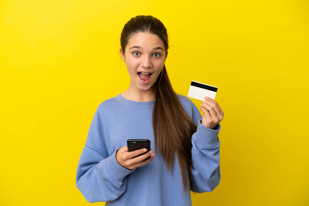 Küçük kız sarı arka planda cep telefonuyla alışveriş yapıyor ve elinde şaşırmış bir ifadeyle bir kredi kartı tutuyor. - Fotoğraf, Görsel
