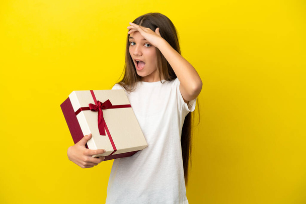 Ragazzina che tiene un regalo su sfondo giallo isolato facendo gesto a sorpresa mentre guarda di lato - Foto, immagini
