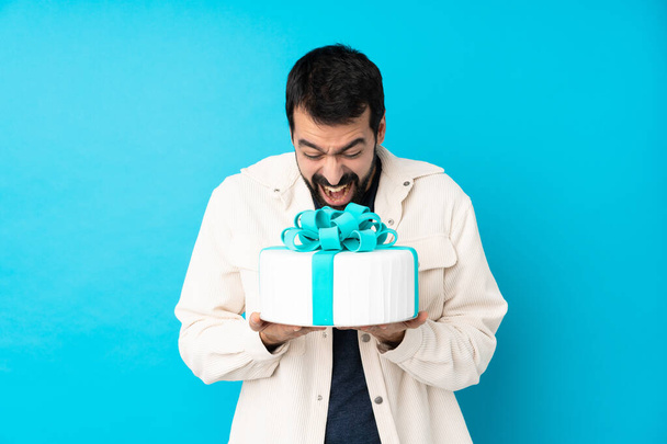Young handsome man with a big cake over isolated blue background - Zdjęcie, obraz