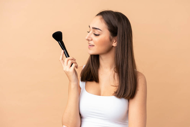 Young caucasian woman isolated on beige background holding makeup brush - Фото, изображение