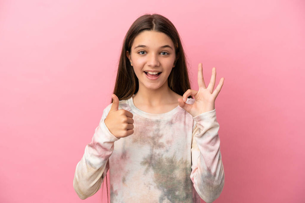Маленькая девочка на изолированном розовом фоне показывает ОК знак и большой палец вверх жеста - Фото, изображение