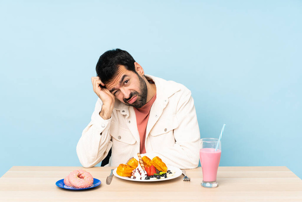 Mężczyzna przy stole je śniadaniowe gofry i shake 'a z wyrazem frustracji i niezrozumienia - Zdjęcie, obraz