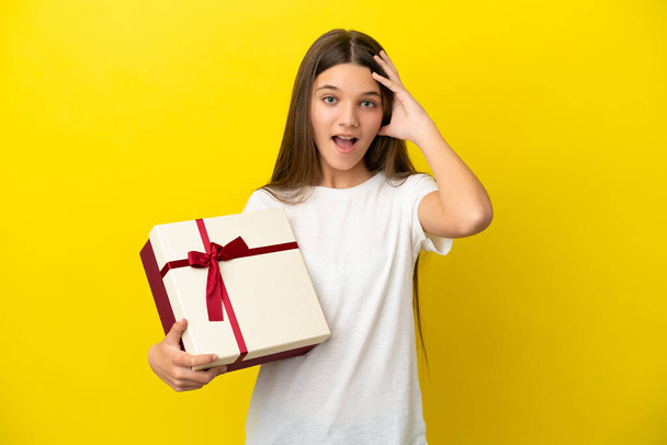 Bambina che tiene un regalo su sfondo giallo isolato con espressione a sorpresa - Foto, immagini