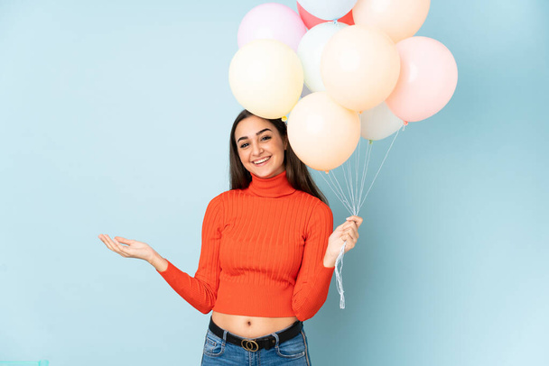 Young woman catching many balloons isolated on blue background holding copyspace imaginary on the palm - Fotografie, Obrázek