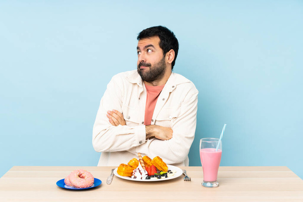 Mężczyzna przy stole jedzący śniadaniowe gofry i shake 'a budzący wątpliwości w geście uniesienia ramion - Zdjęcie, obraz