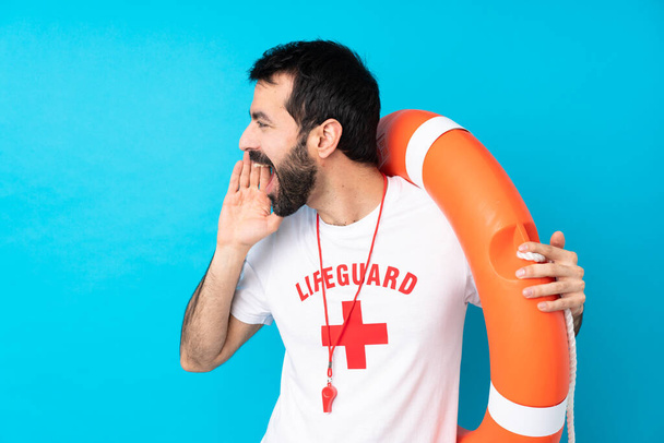 Lifeguard man over isolated blue background shouting with mouth wide open to the lateral - Photo, Image