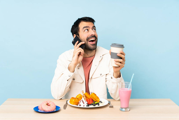 Man at a table having breakfast waffles and a milkshake holding coffee to take away and a mobile - Fotó, kép