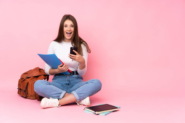 adolescente caucasico studentessa seduta sul pavimento isolato su sfondo rosa sorpreso e l'invio di un messaggio - Foto, immagini