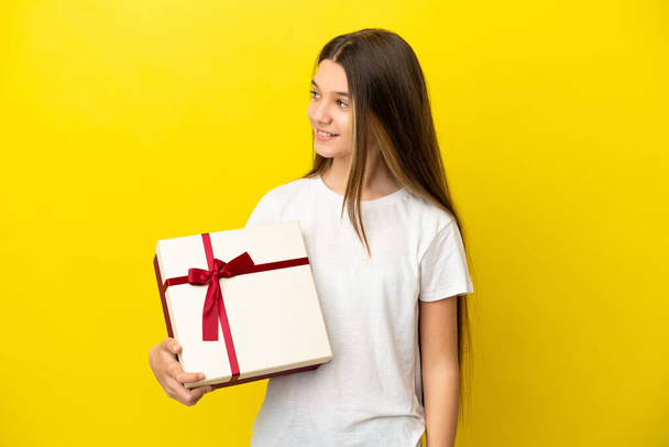 Niña sosteniendo un regalo sobre fondo amarillo aislado mirando lado - Foto, imagen