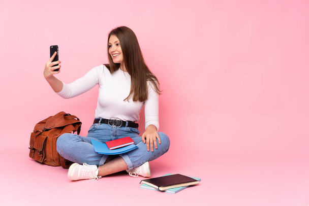 Teenager caucasian student girl sitting on the floor isolated on pink background making a selfie - Photo, Image