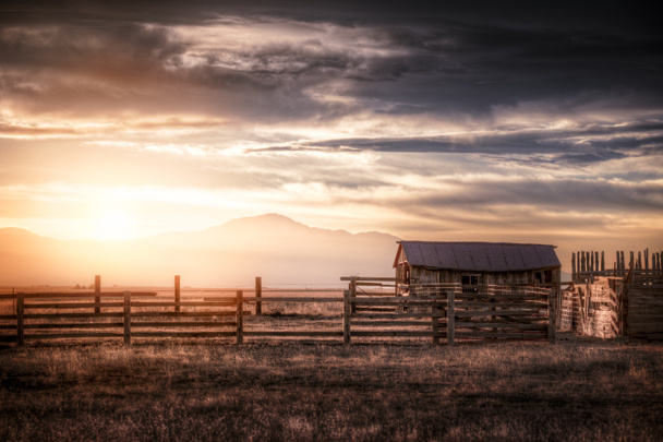 An old farm setting on the rural plains of Colorado under an orange sunset. There is an old fashioned farmhouse or barn along with a corral visible in the scene. It's barren and dry.  - Photo, Image