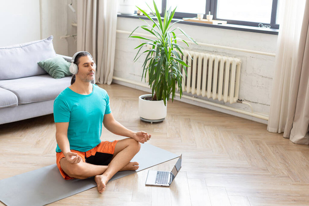 L'entraînement à domicile et le concept de mode de vie sain - l'homme médite à la maison - en utilisant Internet pour surveiller l'entraînement. Il écoute de la musique de méditation relaxante à travers des écouteurs. - Photo, image