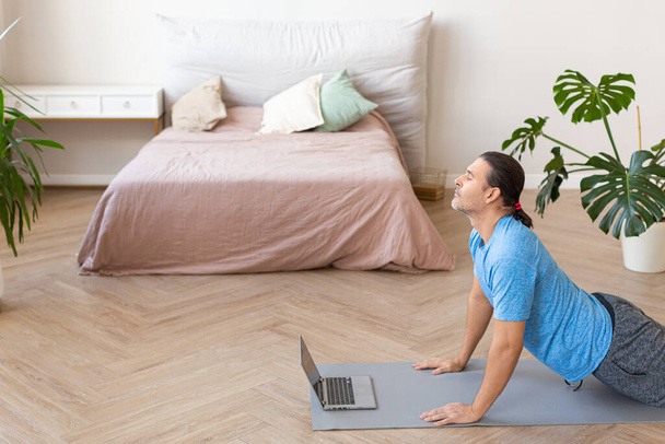 Photo of a man practices pilares at home in front of a laptop monitor. She does a stretching exercise - cobra. - Fotó, kép