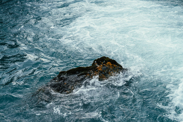 Mossy πέτρα σε γαλάζιο νερό του ποταμού βουνό close-up. Μπλε φόντο της φύσης με ογκόλιθο με βρύα σε τυρκουάζ νερά του ορεινού κολπίσκου. Πλήρες πλαίσιο του θαλάσσιου σερφ. Πέτρα στον ωκεανό surf. Σταγόνα της παλίρροιας - Φωτογραφία, εικόνα