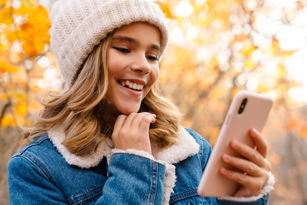 Photo of young happy girl outdoors in autumn park using mobile phone - Foto, immagini