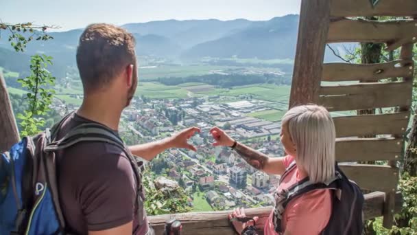 La pareja de excursionistas forma un corazón común con sus dedos en un mirador sobre un pueblo en el valle. Hacer el signo del corazón con las manos en el mirador panorámico. - Metraje, vídeo