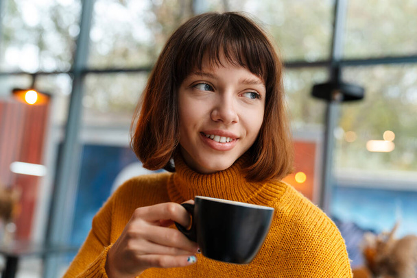 Glückliches rothaariges Mädchen lächelt und trinkt Kaffee, während es im Café drinnen sitzt - Foto, Bild