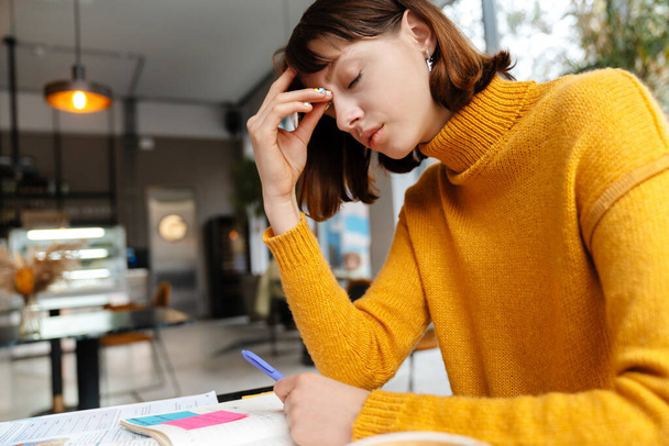 Exhausted redhead student girl with headache doing homework in cafe - Φωτογραφία, εικόνα
