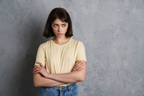 Portrait of an upset young woman standing with arms folded on gray background - 写真・画像