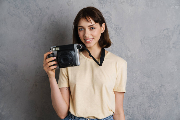 Portrait of a beautiful smiling young girl in casual clothes standing isolated over gray background, holding photo camera - Fotó, kép