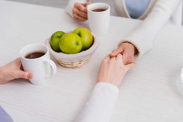 vista recortada de la mujer mayor y la enfermera tomados de la mano cerca de manzanas frescas y tazas de té - Foto, imagen