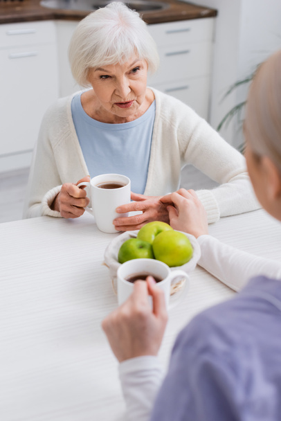 starsza kobieta rozmawia z pracownikiem socjalnym w pobliżu świeżych jabłek i filiżanek herbaty, zamazany pierwszy plan - Zdjęcie, obraz