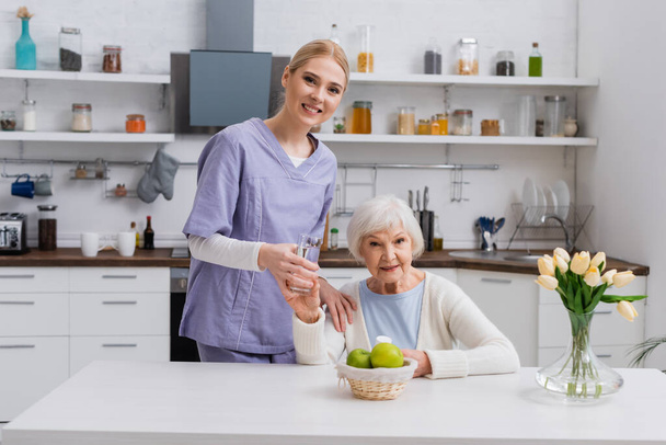 giovane infermiera e anziana donna sorridente alla macchina fotografica mentre tiene in mano un bicchiere d'acqua e farmaci in cucina - Foto, immagini
