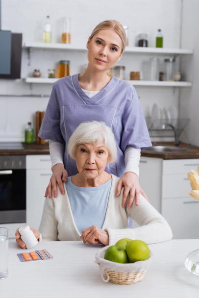 junge Krankenschwester blickt in die Kamera, während sie die Schulter einer Seniorin berührt, die Medikamente in der Hand hält - Foto, Bild