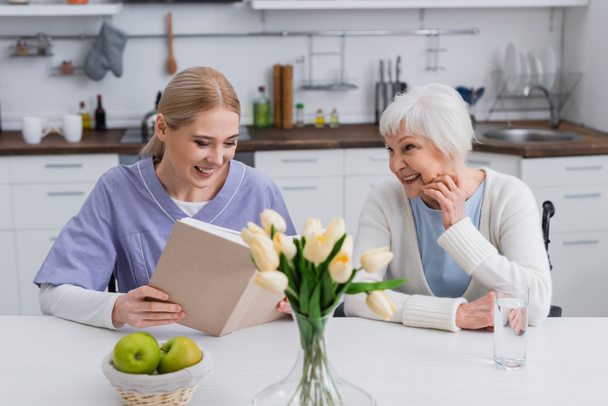 Enfermera sonriente leyendo libro a mujer mayor feliz cerca de manzanas frescas y tulipanes en la cocina - Foto, Imagen