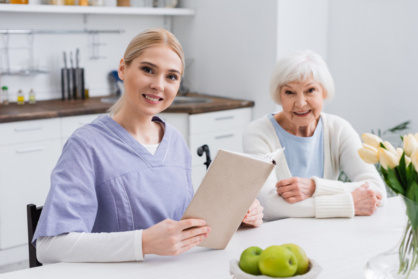 infermiera sorridente con libro guardando la macchina fotografica vicino felice donna anziana in cucina - Foto, immagini
