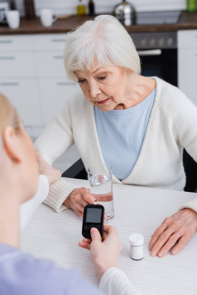 nurse holding glucometer near senior diabetic woman and pills container, blurred foreground - Photo, Image