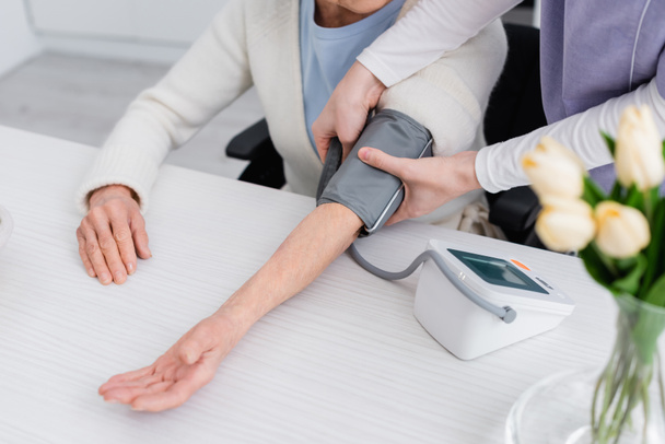 visión parcial de la mujer mayor y enfermera midiendo la presión arterial con tonómetro, primer plano borroso - Foto, Imagen