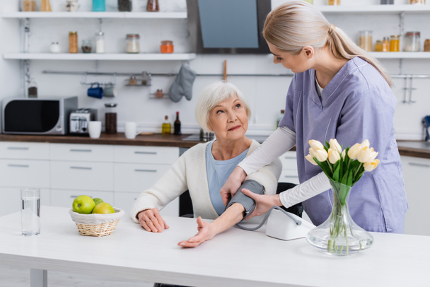 jovem enfermeira e mulher idosa olhando uns para os outros ao medir a pressão arterial na cozinha - Foto, Imagem