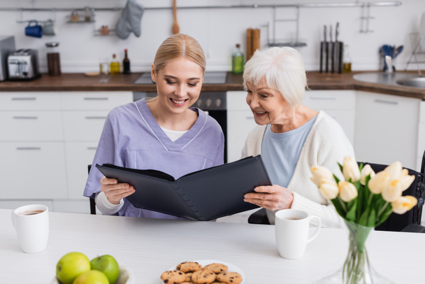 ευχαριστημένη ηλικιωμένη γυναίκα και νοσοκόμα κοιτάζοντας άλμπουμ φωτογραφιών στην κουζίνα - Φωτογραφία, εικόνα
