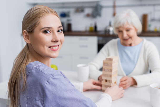 happy nurse looking at camera while playing wooden tower game with blurred senior woman - Foto, Imagem