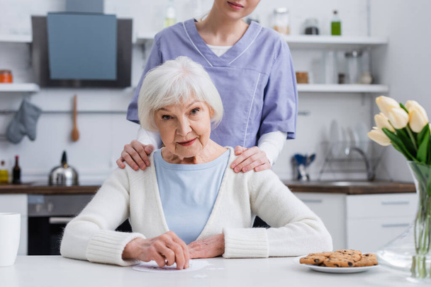 young nurse touching shoulders of elderly woman looking at camera near jigsaw puzzle - Photo, Image