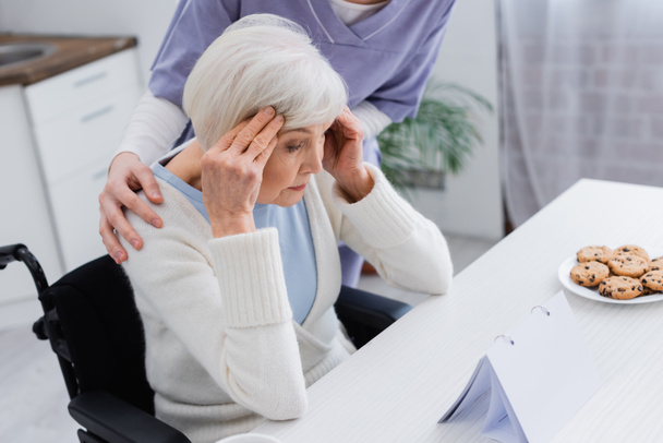 κοινωνική λειτουργός αγκαλιάζει ώμους ηλικιωμένης γυναίκας με ειδικές ανάγκες που υποφέρει από πονοκέφαλο - Φωτογραφία, εικόνα