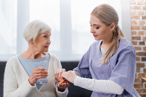 молодая медсестра дает таблетки пожилой женщине держа стакан воды - Фото, изображение