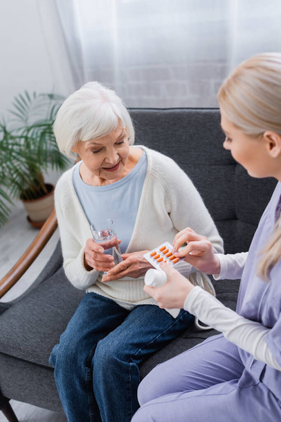 nővér gyógyszert ad idős nő ül a kanapén egy pohár vízzel - Fotó, kép