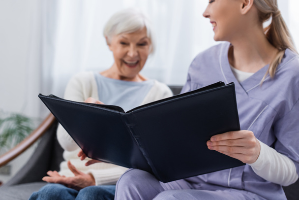 cheerful senior woman with social worker looking at photo album at home, blurred background - Photo, image