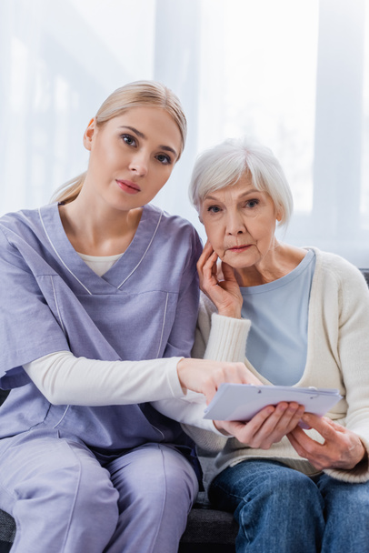 young nurse and senior woman, sick on dementia, looking at camera while holding calendar - Photo, Image