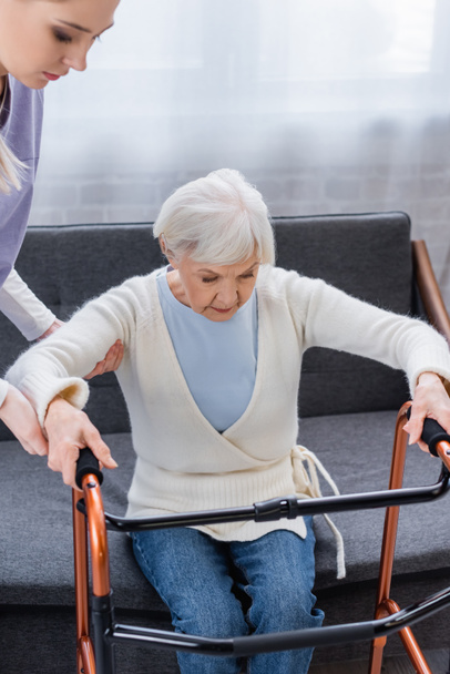 asistente social ayudando a una anciana a levantarse del sofá cerca de caminantes médicos - Foto, Imagen