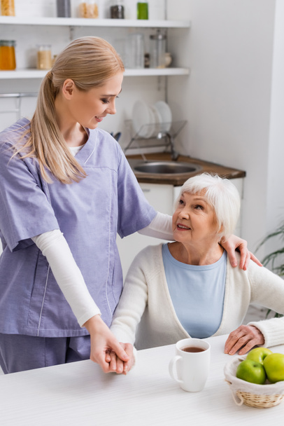 νέα νοσοκόμα κρατώντας το χέρι και αγκαλιάζει τον ώμο της χαρούμενη ηλικιωμένη γυναίκα στην κουζίνα - Φωτογραφία, εικόνα