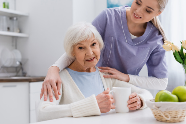 glückliche alte Frau lächelt in die Kamera, während junge Krankenschwester ihre Schultern in der Küche umarmt - Foto, Bild
