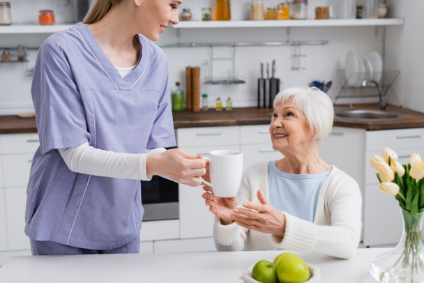 young social worker giving cup of tea to happy elderly woman in kitchen - Foto, afbeelding